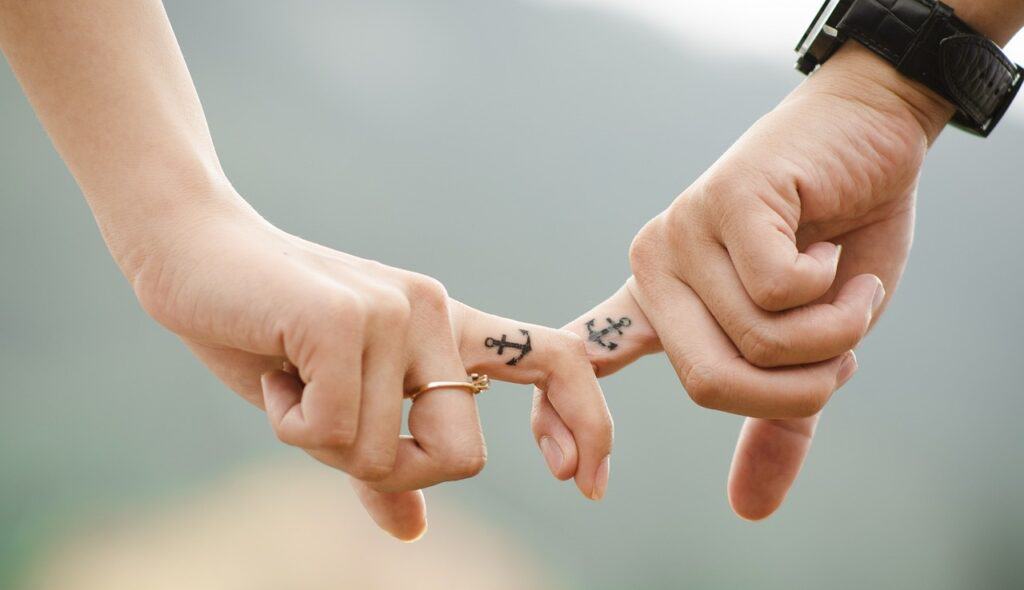Two hands gently holding pinky fingers, demonstrating unhealthy communication in relationships, with anchor tattoos on each, against a blurred natural background.