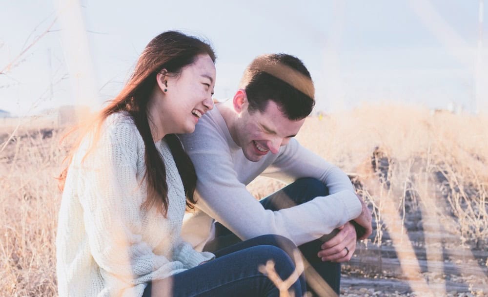 A joyful young couple laughing together while sitting on wooden rails in a grassy field, with unhealthy communication in relationships evident from their soft focus background.