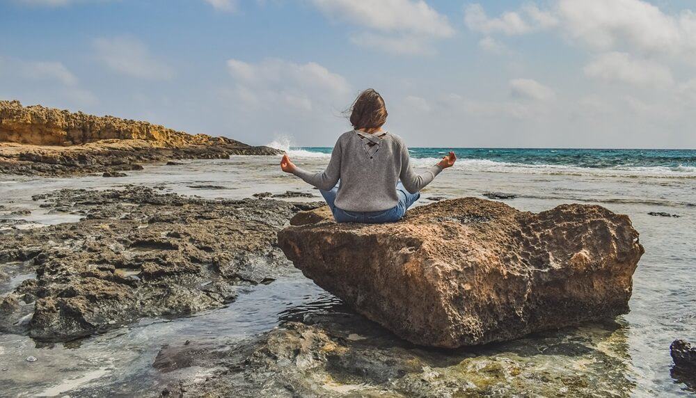 A person practicing healthy lifestyle habits, meditating on a large rock by the sea, with calm water and rocky shore in the background.