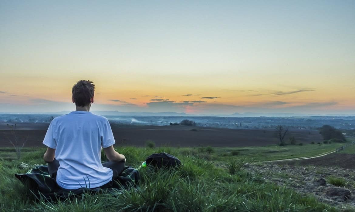 A person practicing healthy lifestyle habits sits cross-legged on a grassy hilltop at dusk, overlooking a panoramic view of a vast landscape with distant mountains and a colorful sky.