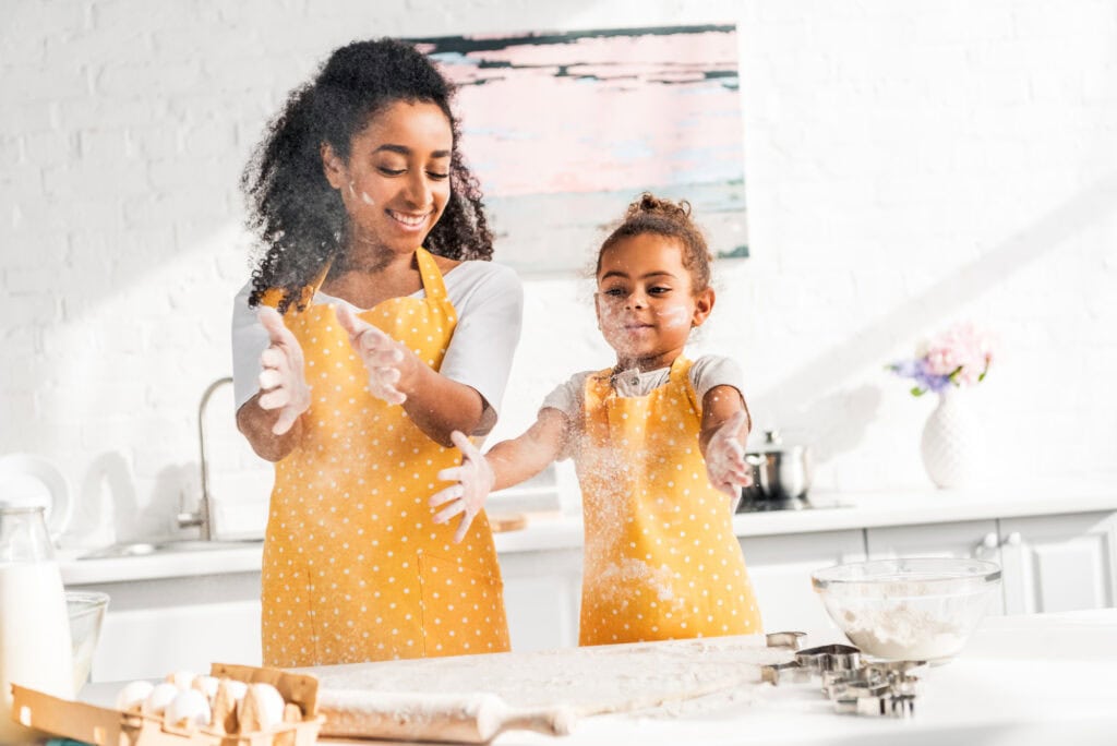 A woman and a child in matching yellow aprons clap their flour-covered hands while baking in a bright kitchen.