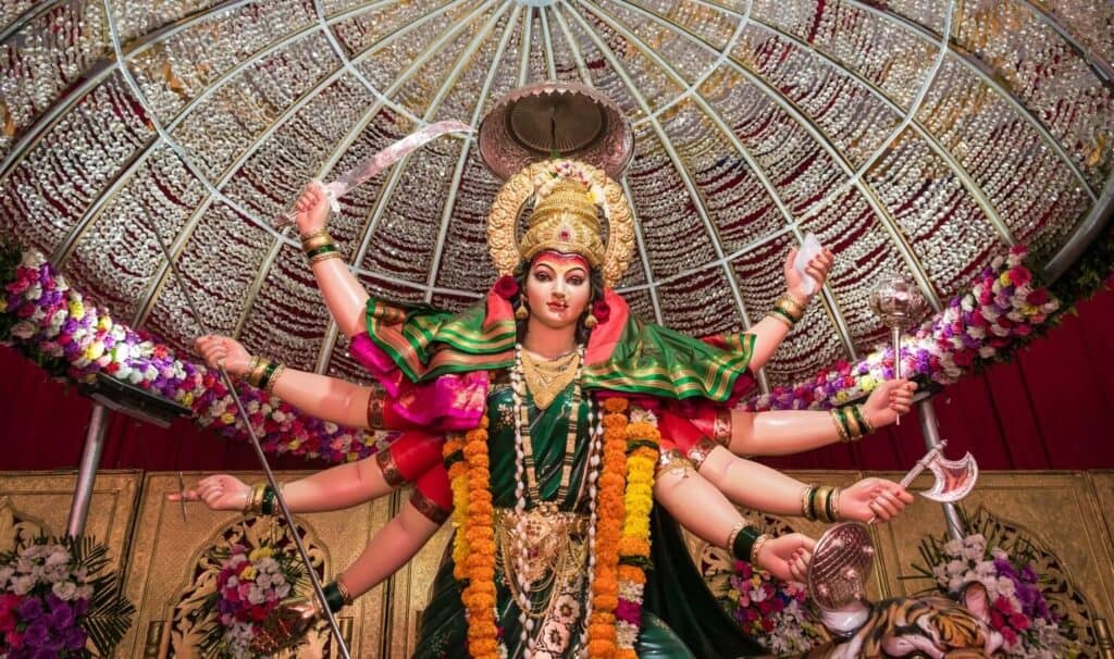 A large statue of a multi-armed deity adorned with colorful flowers and traditional attire, standing under a decorative canopy. Flowers and offerings surround the statue.