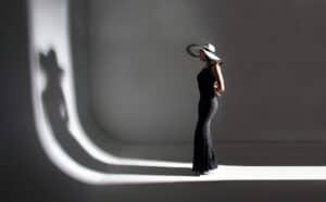 A woman in a long black dress and a wide-brimmed hat stands in profile with her hands behind her back, casting a shadow of her shadow self on a curved white background.