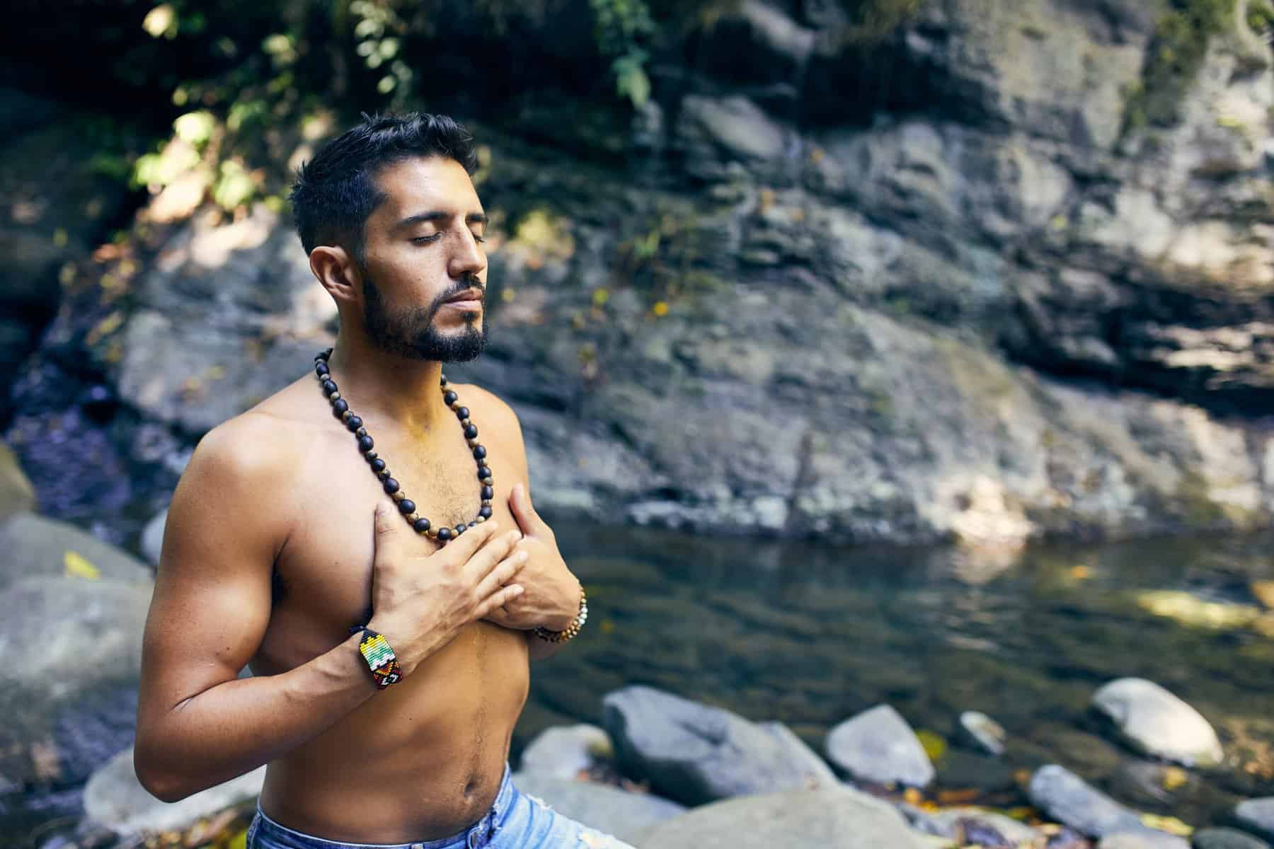 A man stands shirtless near a rocky stream, eyes closed, hands on chest, wearing beaded jewelry and wristbands, embodying spiritual coaching amidst serene natural scenery.