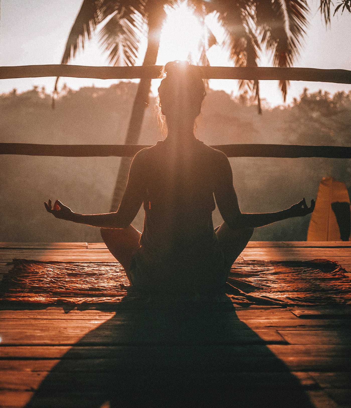 A person sits cross-legged on a wooden deck, meditating at sunrise as part of their spiritual coaching journey. Palm trees are visible in the background.