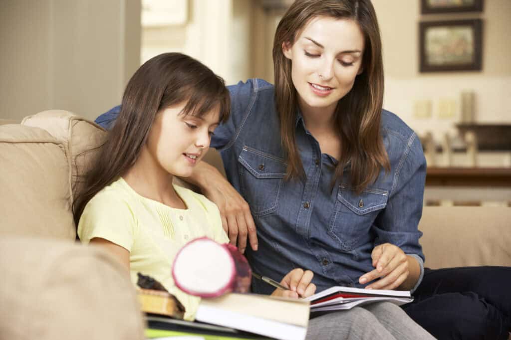 A woman and a young girl sit on a couch. The woman, who is a stepmom, is pointing at a book while the girl follows along, indicating a teaching or homework session filled with support.