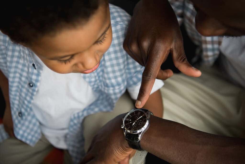 An adult points to a wristwatch on their arm while a child looks on attentively. Both are wearing casual clothing.