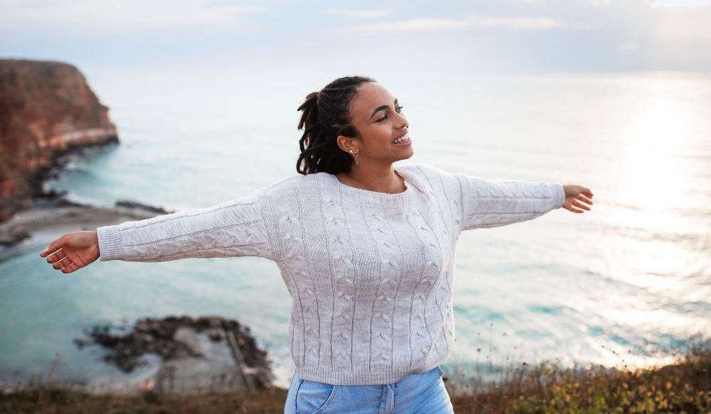 A person, in search of their bliss, stands with arms outstretched near a cliff overlooking the ocean, wearing a white cable-knit sweater and jeans.