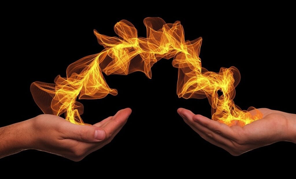 Two hands holding and transferring a swirling, glowing orange flame against a black background (chiron wound)