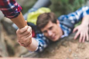 A person in a plaid shirt holds onto another person's hand for support while climbing a rocky surface.