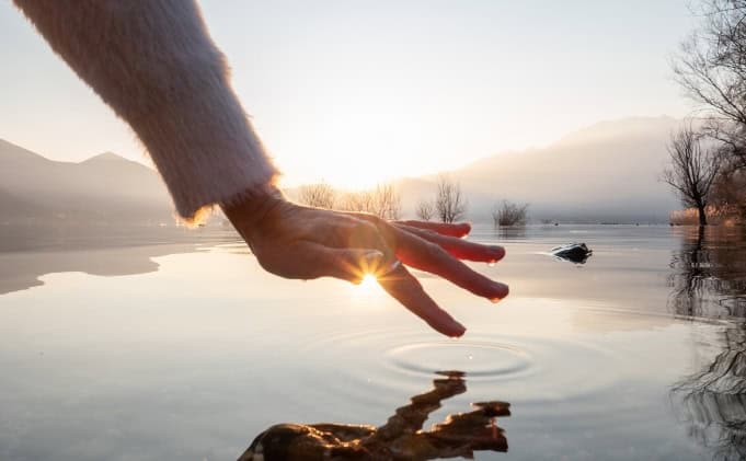 A hand reaching towards the calm surface of a lake at sunrise, with mountains and trees in the background.