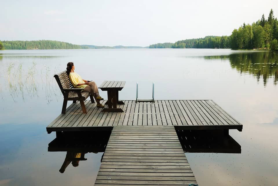 Susan Cain sits on a dock bench in her yellow shirt, gazing thoughtfully at a calm lake surrounded by trees under a clear sky.