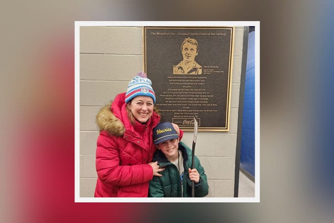 A woman and child in coats and hats pose in front of a commemorative plaque on a wall.