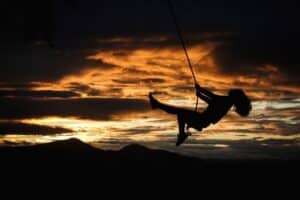 Silhouette of a person swinging on a rope, embracing the shift from FOMO to JOMO against a dramatic, colorful sunset sky with hills in the background.