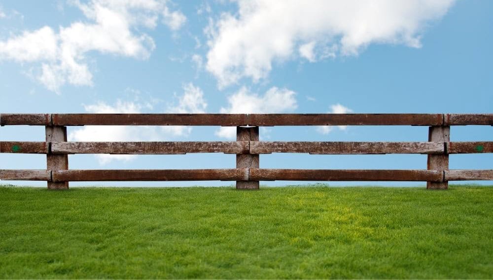A wooden fence stands on a green grassy field under a blue sky with scattered white clouds.