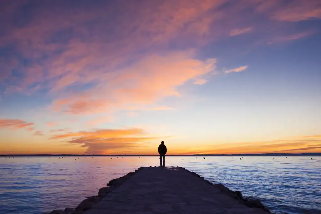A person stands alone at the end of a stone pier overlooking a calm body of water during a colorful sunset with pink and orange clouds.