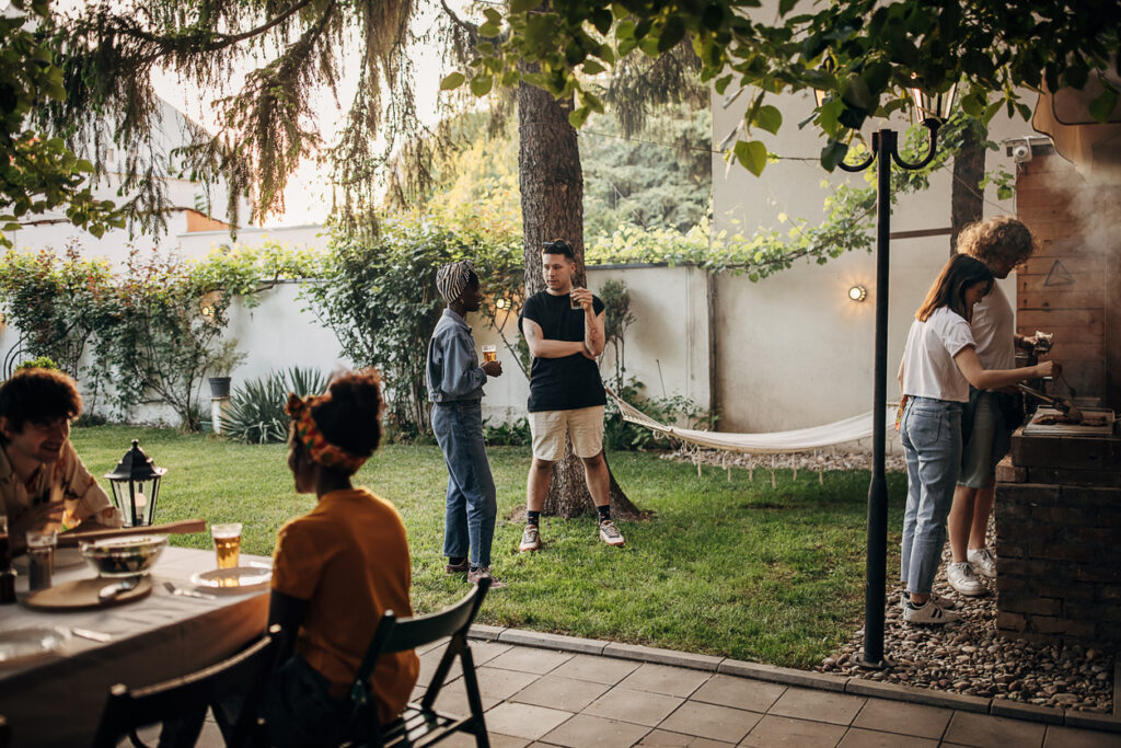 People are gathered in a backyard, some sitting at a table, others standing and talking about self-forgiveness, while two people grill food near a barbecue.