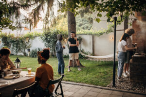 People are gathered in a backyard, some sitting at a table, others standing and talking about self-forgiveness, while two people grill food near a barbecue.