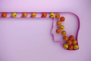 Outline of a human head with smiling, neutral, and frowning emoji balls entering the head through a channel on a pink background, symbolizing different emotions as explored in the Fascinate Test.