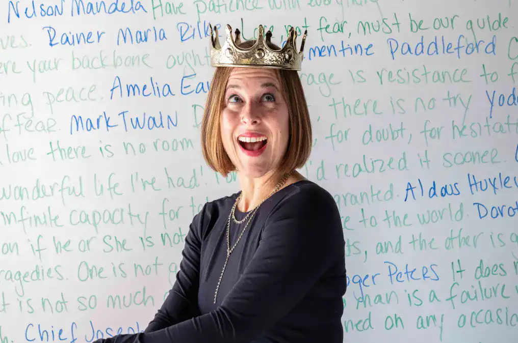 Woman wearing a gold crown stands smiling in front of a whiteboard covered in colorful handwritten quotes and names.