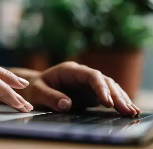 Close-up of a person’s hands typing on a laptop keyboard with a blurred plant in the background.