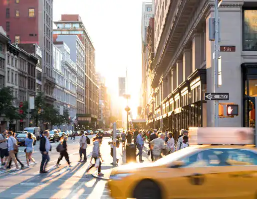 A busy city street at sunset with people crossing the intersection and a yellow taxi passing by in the foreground.