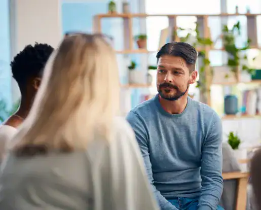 A man in a blue sweater sits in a group setting, listening attentively to others, with shelves of plants visible in the background.