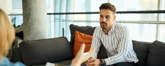 A man sits on a couch attentively listening to another person whose hands are visible in the foreground, suggesting a conversation or interview.
