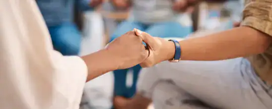 Two people hold hands while sitting in a group, suggesting support or solidarity. Other people are visible in the blurred background.