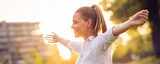 A woman stands outdoors with her arms outstretched, smiling, as sunlight shines in the background.