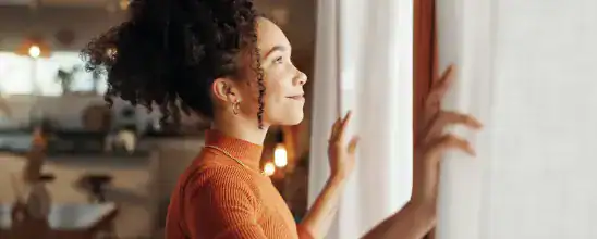A woman with curly hair wearing an orange top stands by a window, gently opening white curtains and looking outside with a slight smile.