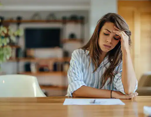 A woman sits at a table with her head resting on her hand, looking stressed while reviewing papers in a home setting.