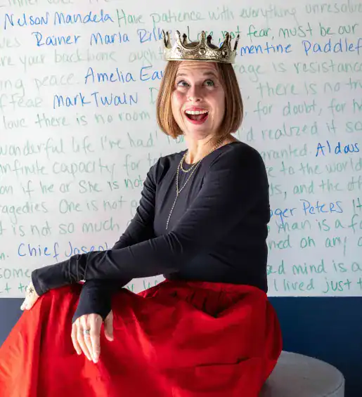 A woman wearing a gold crown, black top, and red skirt sits smiling in front of a wall covered with handwritten quotes.