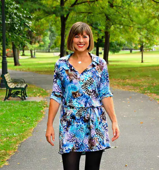 A woman wearing a blue patterned dress and black tights walks on a paved path in a park with green trees and grass.