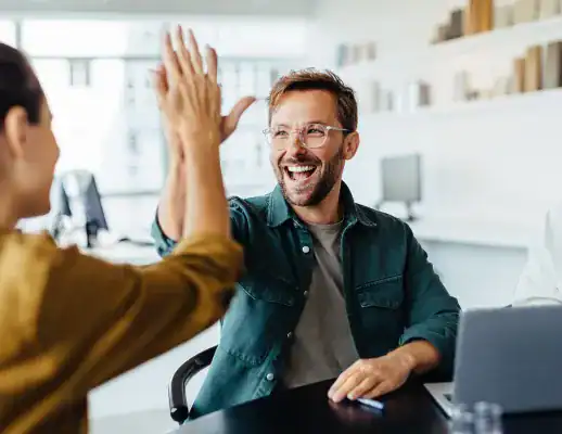 A man and a woman give each other a high five while sitting at a desk with a laptop, smiling in a modern office setting.