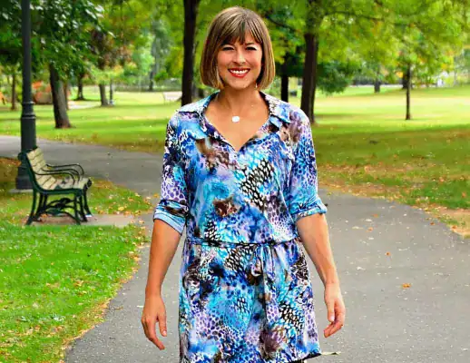 A person with short brown hair, wearing a blue patterned dress, walks on a paved path in a park with trees and benches.