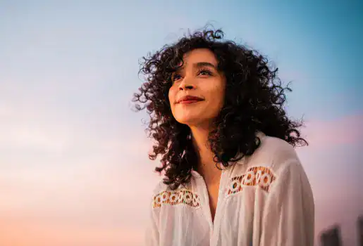 A woman with curly hair and a white blouse looks up and smiles against a colorful sunset sky.