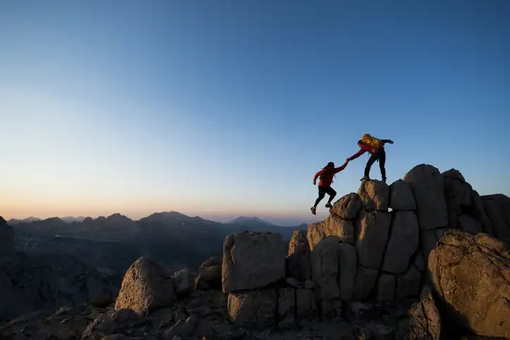 Two hikers on rocky terrain at sunset, with one reaching to help the other climb up a large boulder. Mountain landscape and clear sky in the background.