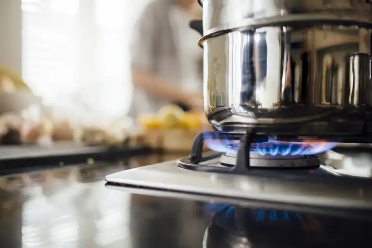 A stainless steel pot sits on a lit gas stove burner, with a blurred person and kitchen counter visible in the background.