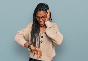 A woman with braided hair and glasses looks worriedly at her watch while touching her head, standing against a plain blue background.