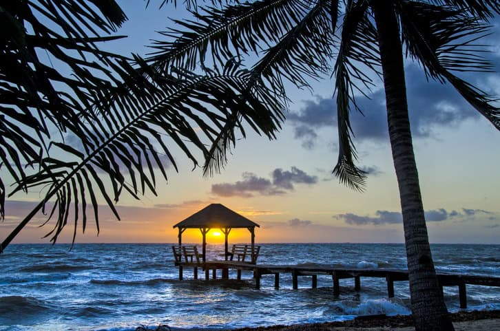 A wooden gazebo on a pier over the ocean at sunset, framed by palm trees and silhouetted against the colorful sky—a true dopamine menu for your senses.
