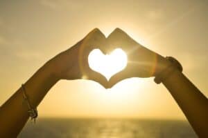 Two hands form a heart shape against a bright sun, with a bracelet visible on one wrist and the ocean in the background.