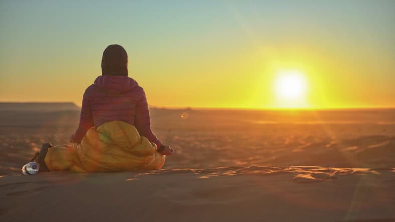 Person sitting on sand in a sleeping bag at sunrise or sunset in a desert landscape, facing the sun. A water bottle is placed beside them.