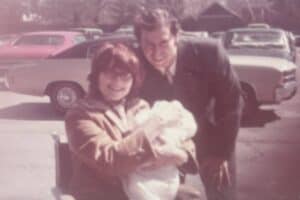 A woman holding a baby sits in a wheelchair next to a smiling man in a suit, with cars parked in the background.